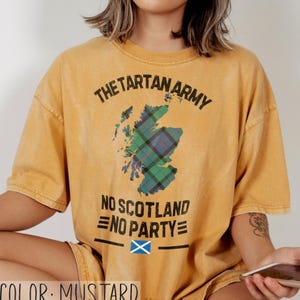 May include: A mustard yellow short-sleeve t-shirt featuring a graphic of the map of Scotland filled with a green, blue, and red tartan pattern. The text reads THE TARTAN ARMY, NO SCOTLAND NO PARTY, and includes a small blue and white Scottish flag.