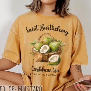 K&ouml;nnte beinhalten: Senffarbenes T-Shirt mit einer Grafik eines Korbes voller gr&uuml;ner Kokosn&uuml;sse und Palmenbl&auml;tter. Der Text auf dem Shirt lautet "Saint Barth&eacute;lemy" und "Caribbean Sea" mit Koordinaten. Das Shirt hat eine lockere Passform.