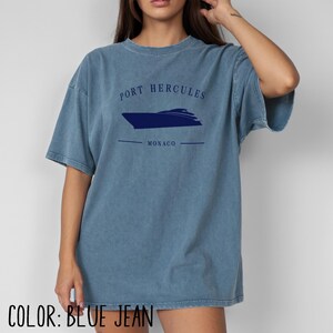 May include: Blue jean-colored t-shirt with the text "PORT HERCULES MONACO" and a navy blue boat graphic. The shirt is a relaxed fit and has short sleeves. The color is a faded, vintage wash.
