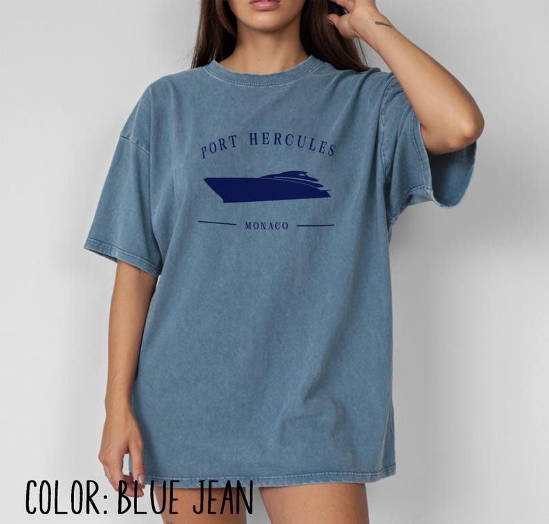 May include: Blue jean-colored t-shirt with the text "PORT HERCULES MONACO" and a navy blue boat graphic. The shirt is a relaxed fit and has short sleeves. The color is a faded, vintage wash.