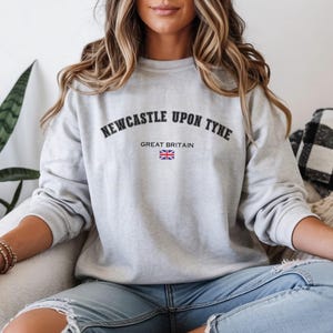 May include: A light gray sweatshirt with the text "NEWCASTLE UPON TYNE" arched across the top and "GREAT BRITAIN" below, with a small Union Jack flag. The sweatshirt is a crew neck style.
