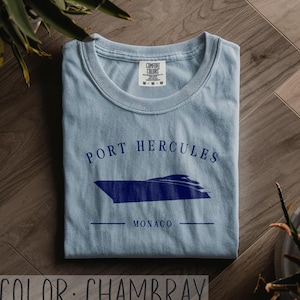 May include: A light blue chambray t-shirt with a navy blue graphic of a boat and the words "PORT HERCULES MONACO". The shirt is folded and has a crew neck. The Comfort Colors tag is visible.