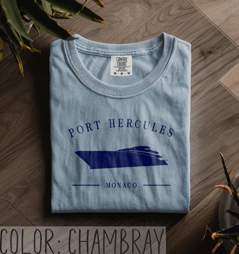 May include: A light blue chambray t-shirt with a navy blue graphic of a boat and the words "PORT HERCULES MONACO". The shirt is folded and has a crew neck. The Comfort Colors tag is visible.