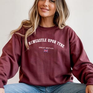 May include: Burgundy sweatshirt with the text "Newcastle Upon Tyne" in white arched lettering, with "Great Britain" and a Union Jack flag underneath. The sweatshirt has long sleeves and a crew neck.