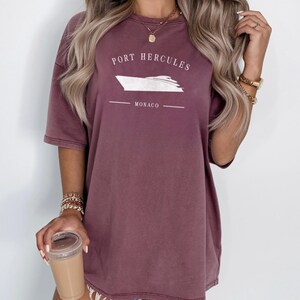 May include: A mauve-colored t-shirt with the text "PORT HERCULES" above a white boat graphic and the word "MONACO" below. The shirt has short sleeves and a relaxed fit.
