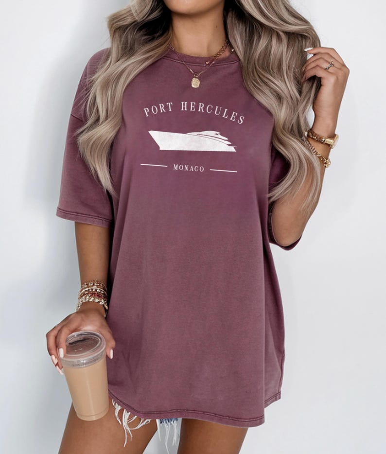 May include: A mauve-colored t-shirt with the text "PORT HERCULES" above a white boat graphic and the word "MONACO" below. The shirt has short sleeves and a relaxed fit.