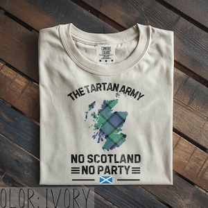 May include: An ivory short-sleeve t-shirt featuring a graphic of the map of Scotland filled with a green and blue tartan pattern. The text reads THE TARTAN ARMY, NO SCOTLAND NO PARTY, and includes a small blue and white Scottish flag icon.