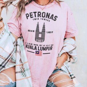 Peut inclure: Un t-shirt rose clair avec un graphique noir des tours Petronas &agrave; Kuala Lumpur. Le texte indique PETRONAS TWIN TOWERS, 451.9 M, 1,483 FT, 3 9 28.08 North, 101 42 42.12 East, KUALA LUMPUR, et comprend un petit drapeau de la Malaisie.