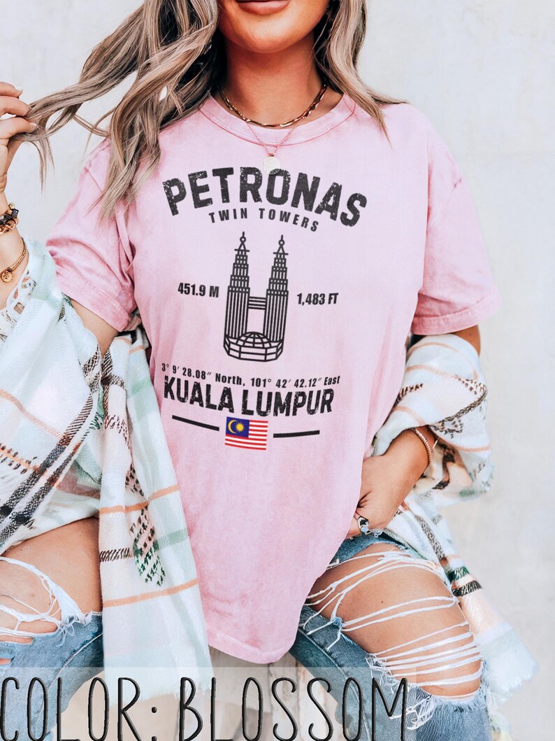 Peut inclure: Un t-shirt rose clair avec un graphique noir des tours Petronas &agrave; Kuala Lumpur. Le texte indique PETRONAS TWIN TOWERS, 451.9 M, 1,483 FT, 3 9 28.08 North, 101 42 42.12 East, KUALA LUMPUR, et comprend un petit drapeau de la Malaisie.