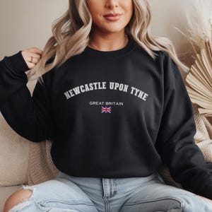 May include: Black crew neck sweatshirt with the text "NEWCASTLE UPON TYNE" in white arched lettering. Below the text is "GREAT BRITAIN" and a small Union Jack flag. The sweatshirt is a solid black color.