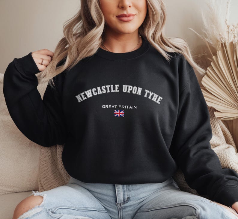 May include: Black crew neck sweatshirt with the text "NEWCASTLE UPON TYNE" in white arched lettering. Below the text is "GREAT BRITAIN" and a small Union Jack flag. The sweatshirt is a solid black color.