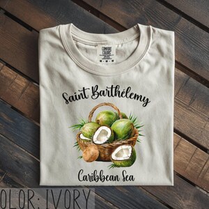 K&ouml;nnte beinhalten: Elfenbeinfarbenes T-Shirt mit einer Aquarellillustration eines Korbes mit gr&uuml;nen Kokosn&uuml;ssen und dem Text "Saint Barth&eacute;lemy Caribbean Sea". Das T-Shirt hat einen Rundhalsausschnitt und ein "Comfort Colors"-Etikett.