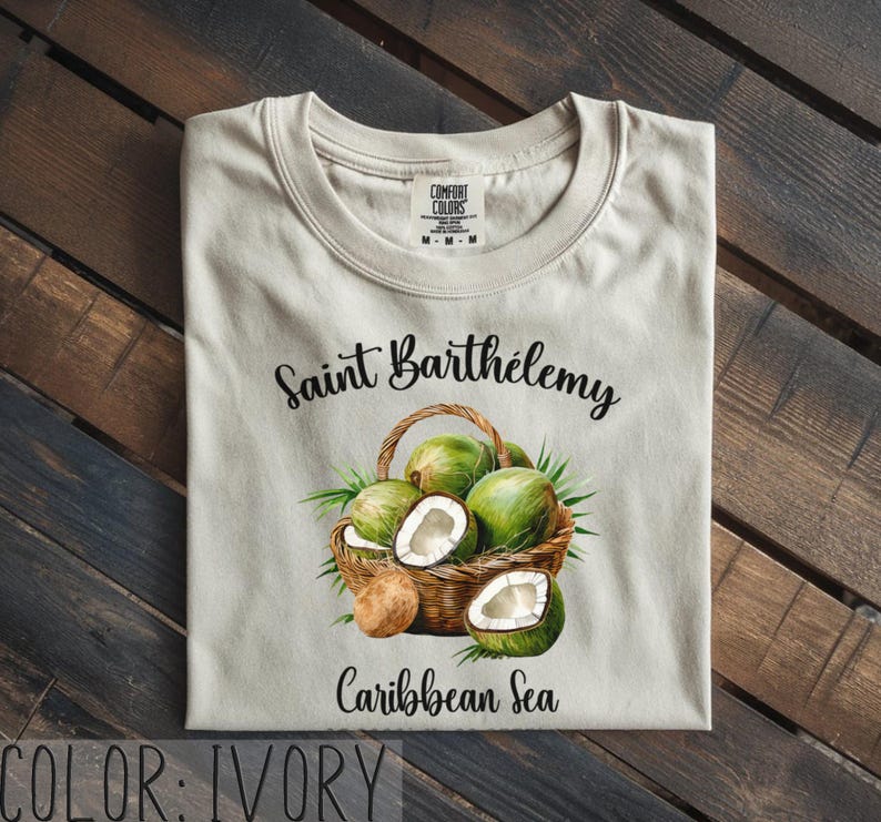 K&ouml;nnte beinhalten: Elfenbeinfarbenes T-Shirt mit einer Aquarellillustration eines Korbes mit gr&uuml;nen Kokosn&uuml;ssen und dem Text "Saint Barth&eacute;lemy Caribbean Sea". Das T-Shirt hat einen Rundhalsausschnitt und ein "Comfort Colors"-Etikett.