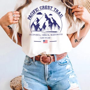 Op de afbeelding: Cr&egrave;mekleurig T-shirt met een blauwe afbeelding van bergen, bomen en de tekst "PACIFIC CREST TRAIL". Daaronder de woorden "CALIFORNIA, OREGON, WASHINGTON - 2.653 MILES -" en een kleine Amerikaanse vlag. De persoon draagt een blauwe spijkerbroekshort en een bruine riem.