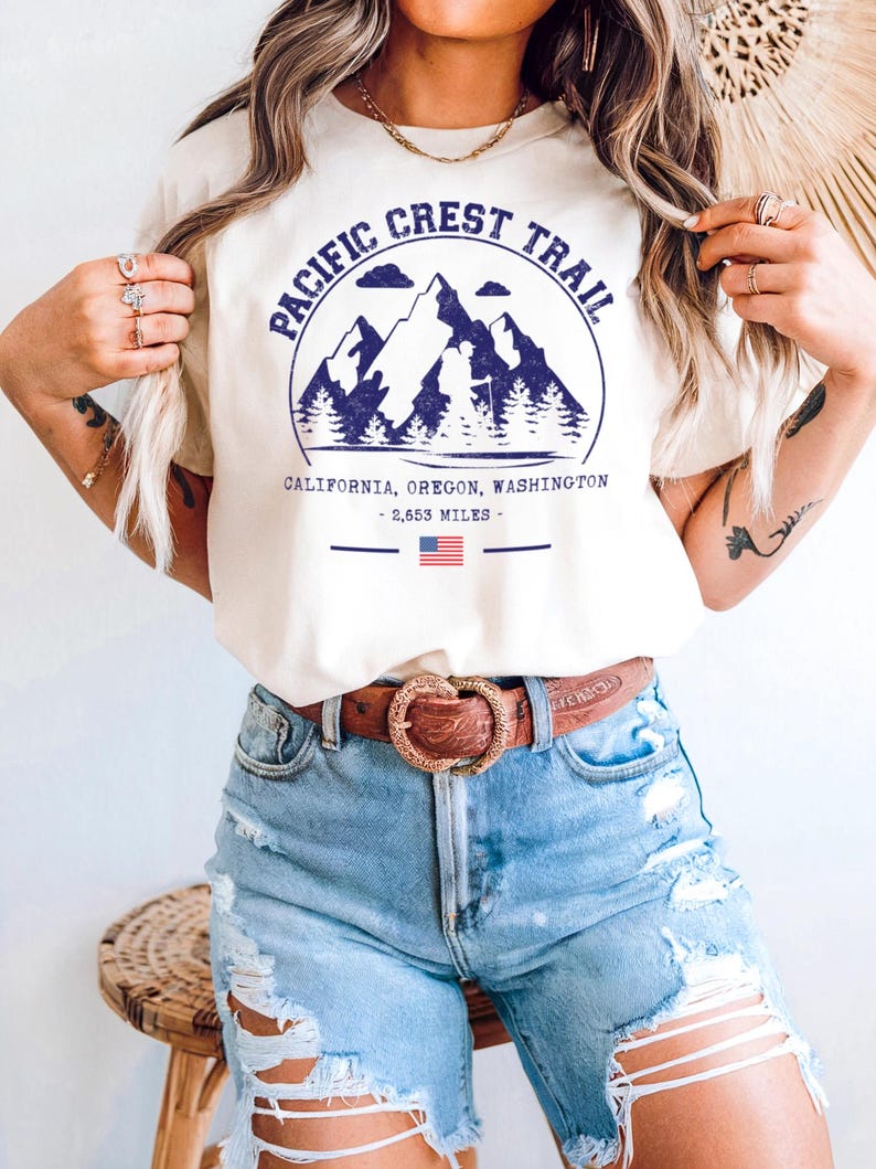 Op de afbeelding: Cr&egrave;mekleurig T-shirt met een blauwe afbeelding van bergen, bomen en de tekst "PACIFIC CREST TRAIL". Daaronder de woorden "CALIFORNIA, OREGON, WASHINGTON - 2.653 MILES -" en een kleine Amerikaanse vlag. De persoon draagt een blauwe spijkerbroekshort en een bruine riem.