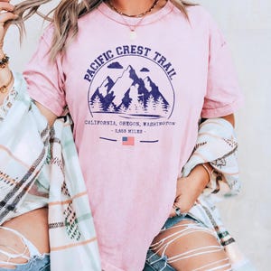 Op de afbeelding: Een lichtroze t-shirt met een cirkelvormige afbeelding met bergen, bomen en de tekst "Pacific Crest Trail". Onder de afbeelding staan de woorden "California, Oregon, Washington" en "2.653 mijl". Er is ook een Amerikaanse vlag aanwezig.