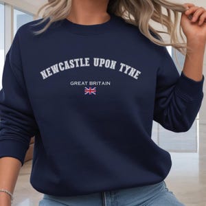 May include: Navy blue sweatshirt with the text "NEWCASTLE UPON TYNE" in white arched lettering. Below the text is "GREAT BRITAIN" and a small Union Jack flag. The sweatshirt is a solid color and has a crew neck.
