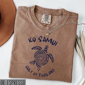 以下が含まれることがあります： エスプレッソカラーの半袖Tシャツ。渦巻き模様の甲羅を持つウミガメのダークブルーのグラフィックがプリントされています。テキストには「Ko Samui」および「Gulf of Thailand」と記されています。クルーネックで、内側にComfort Colorsのブランドラベルが付いています。