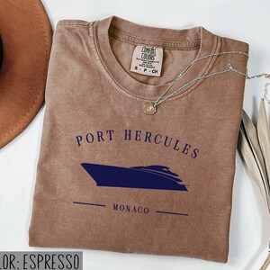 May include: Espresso-colored t-shirt with the text "PORT HERCULES MONACO" and a navy blue boat graphic. The shirt has a crew neck and short sleeves. A silver necklace with a small pendant is draped across the shirt.
