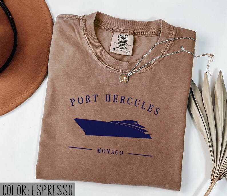 May include: Espresso-colored t-shirt with the text "PORT HERCULES MONACO" and a navy blue boat graphic. The shirt has a crew neck and short sleeves. A silver necklace with a small pendant is draped across the shirt.