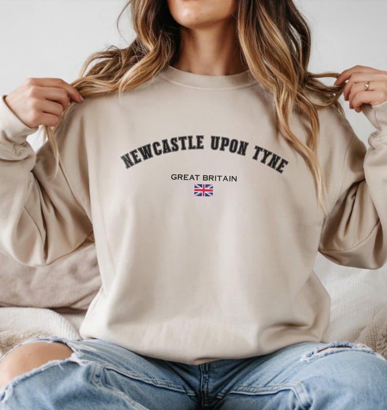 May include: A beige sweatshirt with the text "NEWCASTLE UPON TYNE" in black arched lettering. Below the text is "GREAT BRITAIN" and a small Union Jack flag. The sweatshirt is a neutral color and has long sleeves.