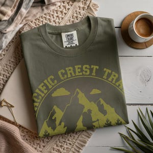 Op de afbeelding: Olijfgroen T-shirt met een ronde hals en de tekst "PACIFIC CREST TRAIL" in een gele, verweerde letter. De afbeelding toont bergen, wolken en een wandelaar. Een kopje koffie staat op een houten onderzetter.