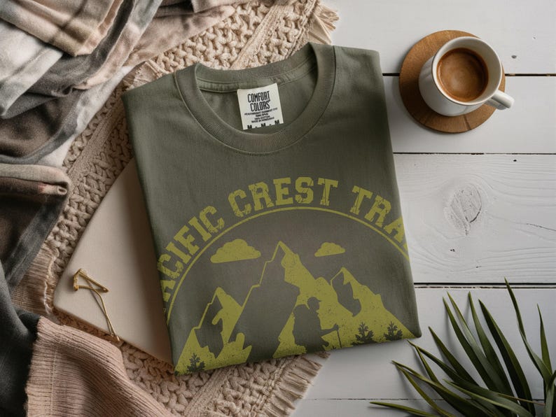 Op de afbeelding: Olijfgroen T-shirt met een ronde hals en de tekst "PACIFIC CREST TRAIL" in een gele, verweerde letter. De afbeelding toont bergen, wolken en een wandelaar. Een kopje koffie staat op een houten onderzetter.