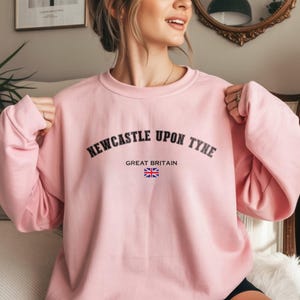 May include: A light pink sweatshirt with the text "NEWCASTLE UPON TYNE" in black arched lettering. Below the text is "GREAT BRITAIN" and a small Union Jack flag. The sweatshirt has long sleeves.