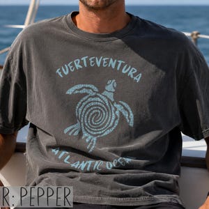 以下が含まれることがあります： ペッパーカラーの半袖Tシャツ。渦巻き模様の甲羅を持つウミガメのライトブルーのグラフィックが特徴です。カメの上部には「FUERTEVENTURA」、カメのグラフィックの下部には「ATLANTIC OCEAN」という文字がプリントされています。