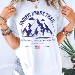 Op de afbeelding: Wit T-shirt met een marineblauwe afbeelding van bergen, bomen en de tekst "Pacific Crest Trail". Onder de afbeelding staan de woorden "California, Oregon, Washington - 2.653 mijl" en een kleine Amerikaanse vlag.