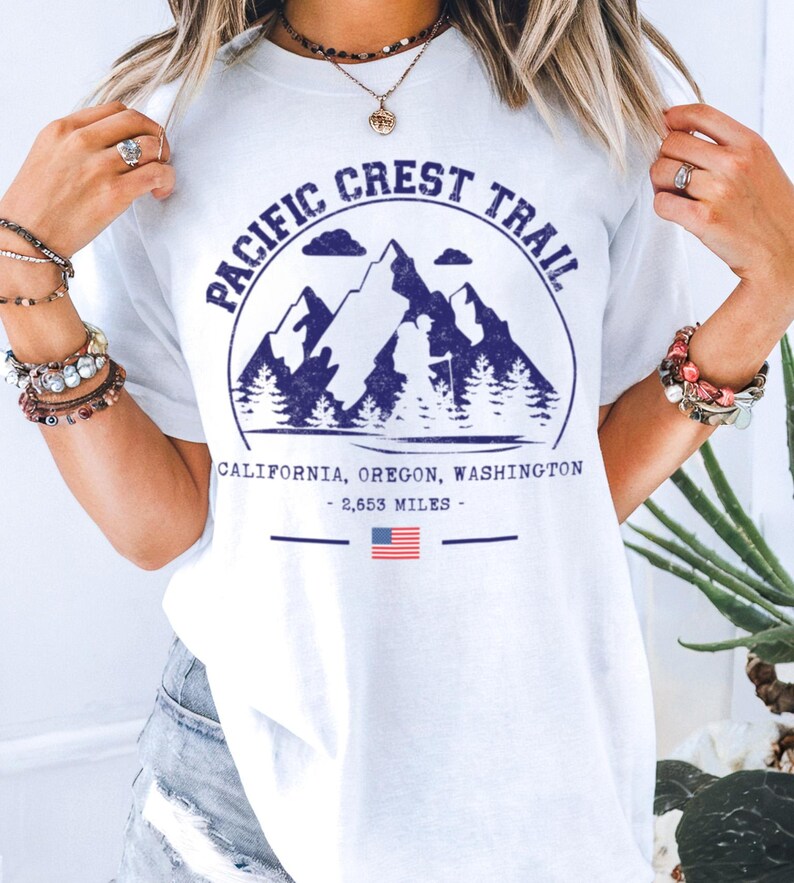 Op de afbeelding: Wit T-shirt met een marineblauwe afbeelding van bergen, bomen en de tekst "Pacific Crest Trail". Onder de afbeelding staan de woorden "California, Oregon, Washington - 2.653 mijl" en een kleine Amerikaanse vlag.