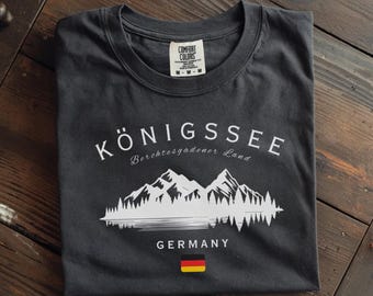 May include: Pepper-colored t-shirt with a white graphic of mountains, trees, and the text "KÖNIGSSEE Berchtesgadener Land GERMANY". The German flag is displayed below the text. The shirt is folded on a wooden surface.
