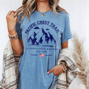 Op de afbeelding: Lichtblauw T-shirt met een marineblauwe afbeelding van bergen, bomen en de tekst "Pacific Crest Trail". De tekst hieronder luidt "California, Oregon, Washington - 4.269 kilometer". Een Amerikaanse vlag is ook afgebeeld.
