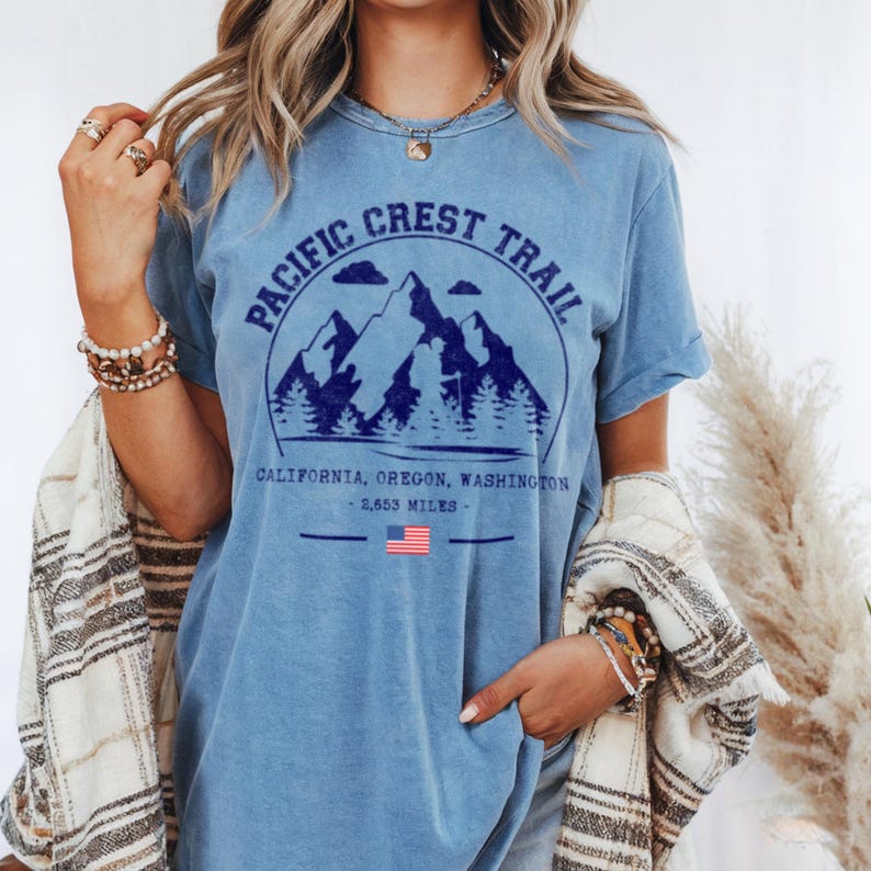 Op de afbeelding: Lichtblauw T-shirt met een marineblauwe afbeelding van bergen, bomen en de tekst "Pacific Crest Trail". De tekst hieronder luidt "California, Oregon, Washington - 4.269 kilometer". Een Amerikaanse vlag is ook afgebeeld.