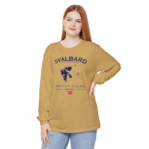 Può includere: Una maglia a maniche lunghe color giallo senape con una grafica blu scuro dell'arcipelago delle Svalbard, una rosa dei venti e il testo Svalbard, Spitzbergen, Arctic Ocean, Norway, accompagnata da una piccola icona della bandiera norvegese.