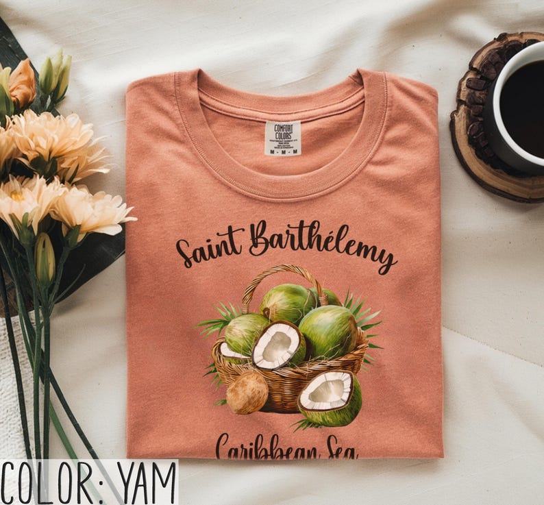 K&ouml;nnte beinhalten: Ein yamfarbenes T-Shirt mit Rundhalsausschnitt. Das Shirt zeigt eine Grafik eines Korbes voller gr&uuml;ner Kokosn&uuml;sse und den Text "Saint Barth&eacute;lemy Caribbean Sea" in schwarzer Schrift. Das Shirt ist auf einer wei&szlig;en Oberfl&auml;che gefaltet.
