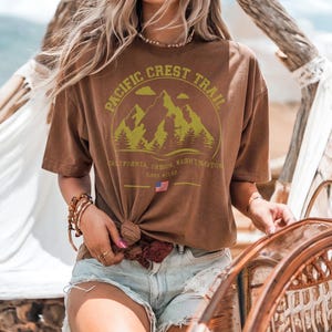 Op de afbeelding: Bruin t-shirt met een bergafbeelding en de tekst "PACIFIC CREST TRAIL". Het t-shirt bevat ook de tekst "CALIFORNIA, OREGON, WASHINGTON" en "2.653 MILES". Het t-shirt is in de taille geknoopt.