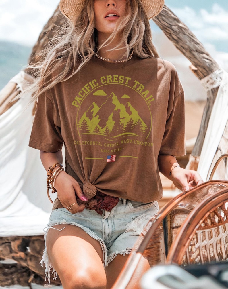 Op de afbeelding: Bruin t-shirt met een bergafbeelding en de tekst "PACIFIC CREST TRAIL". Het t-shirt bevat ook de tekst "CALIFORNIA, OREGON, WASHINGTON" en "2.653 MILES". Het t-shirt is in de taille geknoopt.