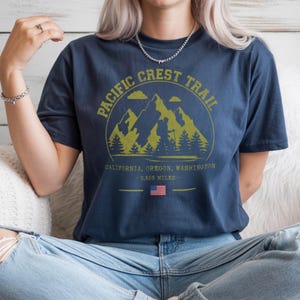 Op de afbeelding: Marineblauw T-shirt met een cirkelvormige afbeelding met bergen, bomen en een wandelaar, met de tekst "PACIFIC CREST TRAIL". Onder de afbeelding staan de woorden "CALIFORNIA, OREGON, WASHINGTON - 2.653 MILES" en een Amerikaanse vlag.