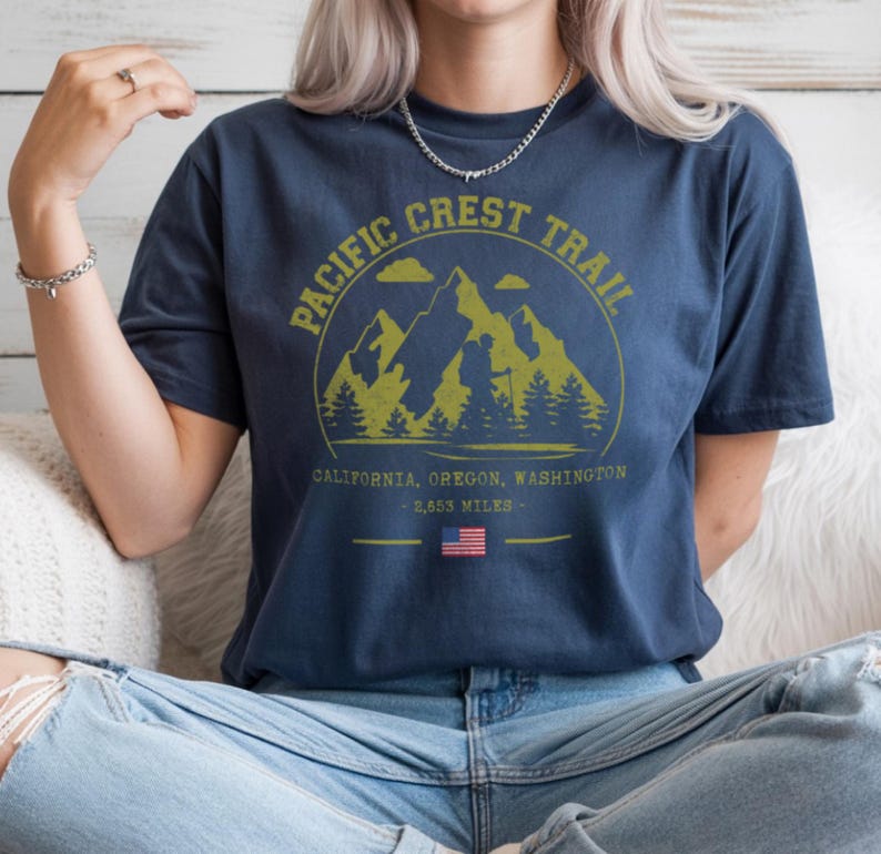 Op de afbeelding: Marineblauw T-shirt met een cirkelvormige afbeelding met bergen, bomen en een wandelaar, met de tekst "PACIFIC CREST TRAIL". Onder de afbeelding staan de woorden "CALIFORNIA, OREGON, WASHINGTON - 2.653 MILES" en een Amerikaanse vlag.