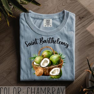 K&ouml;nnte beinhalten: Hellblaues T-Shirt mit dem Text "Saint Barth&eacute;lemy" &uuml;ber einer Aquarellillustration eines Korbes voller gr&uuml;ner Kokosn&uuml;sse und Kokosnussh&auml;lften. Das Shirt ist mit "Comfort Colors" gekennzeichnet. Die Farbe ist "Chambray".