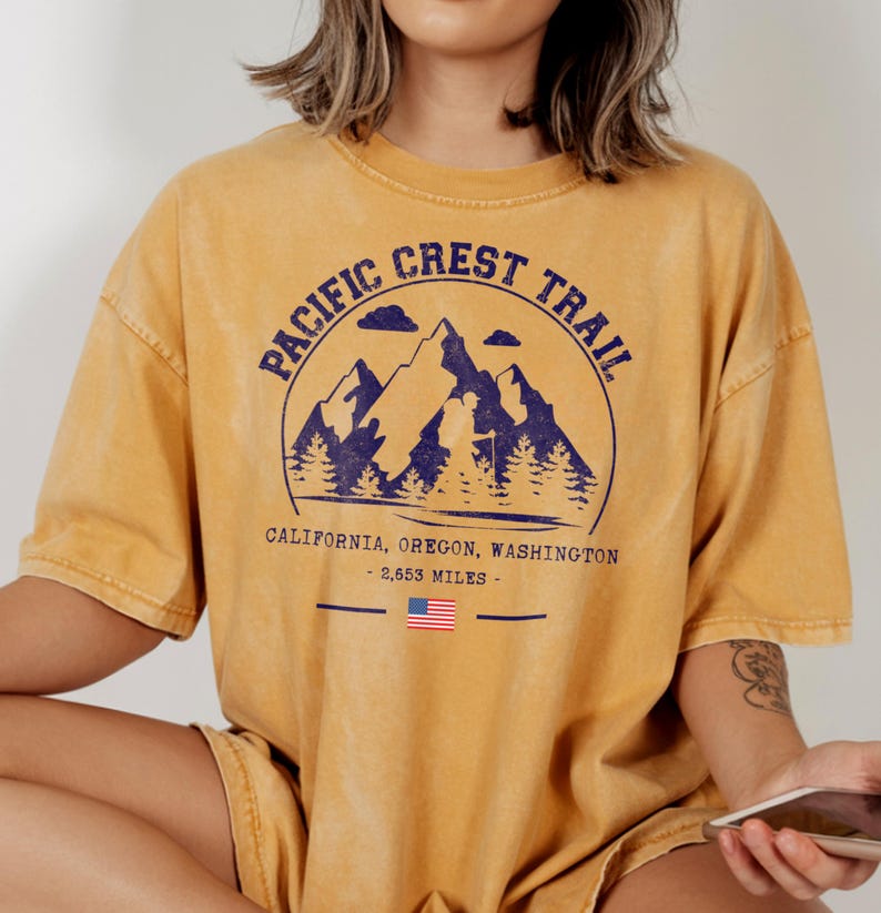 Op de afbeelding: Mosterdgeel T-shirt met een marineblauwe afbeelding van bergen, bomen en de tekst "PACIFIC CREST TRAIL". Onder de afbeelding staan de woorden "CALIFORNIA, OREGON, WASHINGTON - 2.653 MILES" en een Amerikaanse vlag.