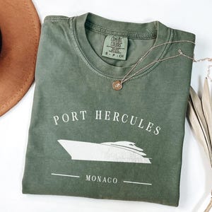 May include: An olive green t-shirt with the words "Port Hercules Monaco" and a boat graphic in white. A gold necklace with a pendant rests on the shirt. A brown hat is visible in the upper left corner.