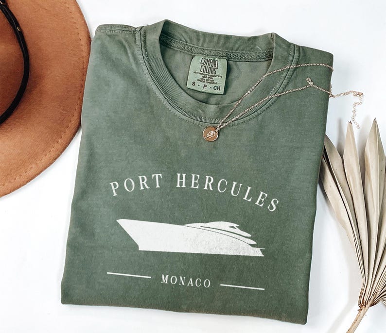 May include: An olive green t-shirt with the words "Port Hercules Monaco" and a boat graphic in white. A gold necklace with a pendant rests on the shirt. A brown hat is visible in the upper left corner.