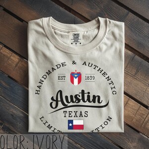 May include: Ivory-colored t-shirt with the text "Austin Texas" in a black script font. The shirt features a circular design with the words "Handmade & Authentic" and the year "1839". A Texas flag is displayed below the text.
