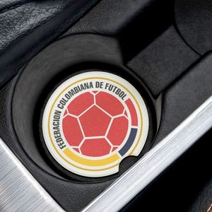 May include: A car cup holder coaster featuring the Colombian Football Federation logo. The coaster has a soccer ball design in red, yellow, and blue, with the text "FEDERACION COLOMBIANA DE FUTBOL" around the edge. The coaster is placed in a car's cup holder.