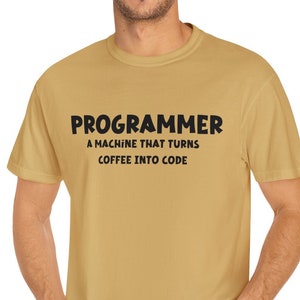Puede incluir: Una camiseta de manga corta de color beige con el texto en negro: PROGRAMMER A MACHINE THAT TURNS COFFEE INTO CODE. La prenda cuenta con un cuello redondo y un corte estándar, ofreciendo un estilo informal en un tono tierra neutro.