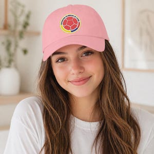 Puede incluir: Gorra de b&eacute;isbol rosa claro con un emblema circular con un dise&ntilde;o de bal&oacute;n de f&uacute;tbol y texto. El emblema es rojo, amarillo y azul. La gorra la lleva una persona con una camiseta blanca.