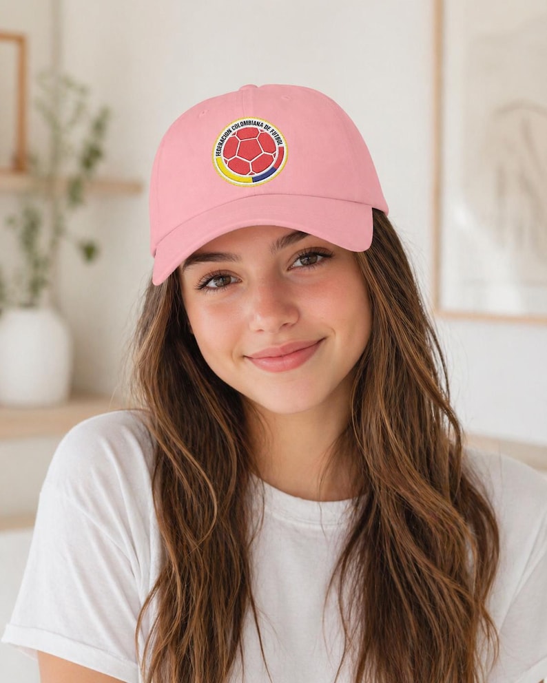 Puede incluir: Gorra de b&eacute;isbol rosa claro con un emblema circular con un dise&ntilde;o de bal&oacute;n de f&uacute;tbol y texto. El emblema es rojo, amarillo y azul. La gorra la lleva una persona con una camiseta blanca.
