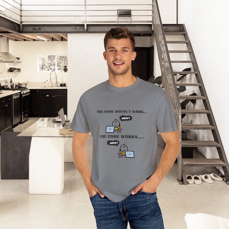 May include: A gray t-shirt with the text "THE CODE DOESN'T WORK... WHY? THE CODE WORKS..... WHY?" and a cartoon graphic of a person at a laptop. The shirt is being worn by a person.
